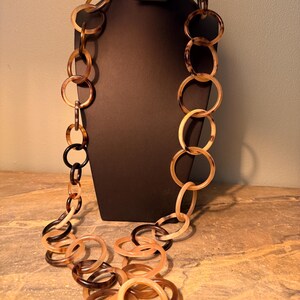 May include: A long necklace composed of numerous interlocking circular rings in varying sizes. The rings feature a translucent, marbled pattern in shades of amber, dark brown, cream, and black, creating a continuous chain-link design.