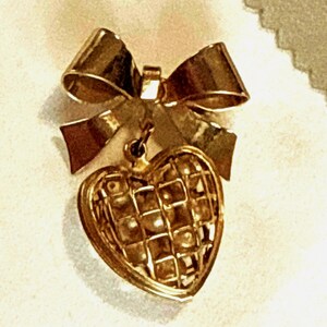 May include: A gold-toned metal brooch featuring a bow at the top with a dangling heart-shaped pendant. The heart pendant has a three-dimensional cage-like structure with a grid pattern, suspended from the center of the bow by a small metal loop.
