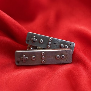 May include: A pair of silver-toned metal cufflinks shaped like rectangular video game remote controllers. Each cufflink features embossed buttons including a plus sign, a letter A, a circular button, a grid pattern, and the numbers one and two.