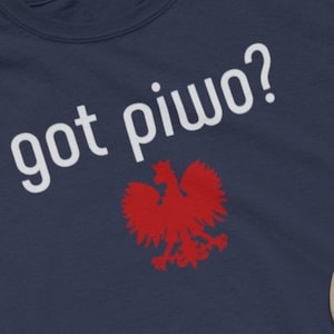 Puede incluir: Una camiseta azul marino con el texto "got piwo?" en blanco y un gráfico de águila polaca roja. La camiseta está colocada sobre una superficie beige con un par de pantalones cortos de mezclilla azul, zapatillas negras, una gorra de béisbol beige y unos auriculares negros.