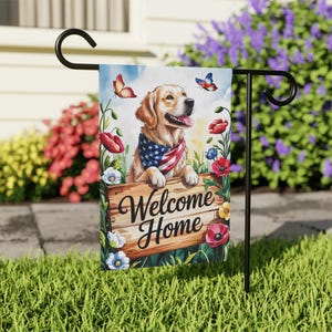 May include: A garden flag with a Golden Retriever wearing an American flag bandana, with the words "Welcome Home" on a wooden sign. The flag is decorated with butterflies and flowers in a garden setting.