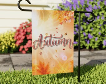 Puede incluir: Una bandera de jardín con la palabra "Autumn" en cursiva, sobre un fondo otoñal cálido con hojas cayendo. La bandera se muestra en un soporte de metal negro, con césped verde y flores coloridas en el fondo.