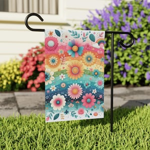 May include: A colorful garden flag featuring a vibrant floral design with a rainbow background. The flag showcases various flowers in shades of pink, orange, blue, and white, with a small rainbow and clouds. The flag is displayed on a black metal stand in a grassy yard.