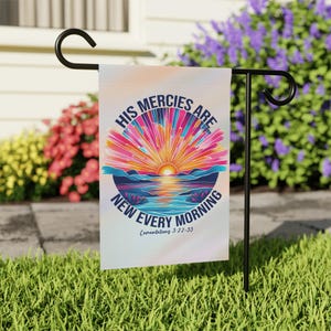 May include: A garden flag displaying the text "HIS MERCIES ARE NEW EVERY MORNING" in a circular design. The flag shows a colourful sunrise over a lake and mountains. The flag is suspended on a black metal stand in a grassy garden.