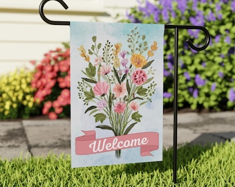 May include: A garden flag featuring a watercolor bouquet of pink, yellow, and orange flowers with green foliage. A pink banner at the bottom reads "Welcome" in white script. The flag is displayed on a black metal stand in a grassy yard.