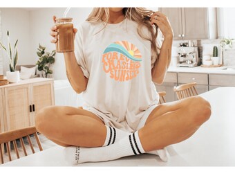 May include: Cream-colored t-shirt with a circular graphic featuring a sunset design and the words "Forever Chasing Sunsets" in orange. The shirt is paired with white socks with black stripes and a glass of iced coffee.