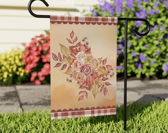Puede incluir: Una bandera de jardín con un diseño floral en acuarela, con rosas y hojas en tonos rosa, melocotón y dorado. Tiene un borde de cuadros marrón y blanco y un borde festoneado. La bandera se muestra en un soporte de metal negro en un jardín cubierto de hierba.