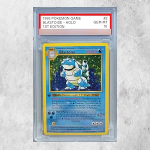 May include: A 1999 Pokemon Game Blastoise Holo 1st Edition trading card graded GEM MT 10, encased in a clear protective plastic slab. The card features the blue Blastoise character, a holographic background, and text detailing its stats and moves.