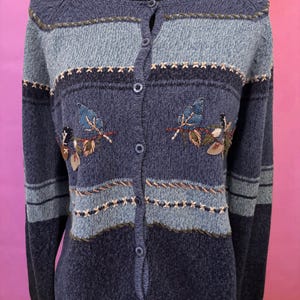 May include: A blue cardigan sweater with a button-up front. The sweater features embroidered leaf and floral designs, along with horizontal stripes in varying shades of blue and tan. The sweater is displayed on a black mannequin.