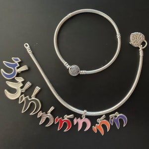 May include: A silver charm bracelet with a heart-shaped charm and several charms with colorful enamel. The charms are shaped like the Hebrew letter 'nun'.