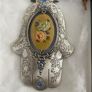 May include: Silver-toned Hamsa hand pendant with an oval center featuring a floral design in yellow, pink, and blue. The pendant is adorned with blue gemstones and a brown leather cord for hanging.