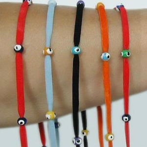 May include: A collection of colorful bracelets, each featuring a cord in red, blue, black, and orange. Each bracelet has a small, round bead with an eye design. The bracelets are worn on a person's arm.
