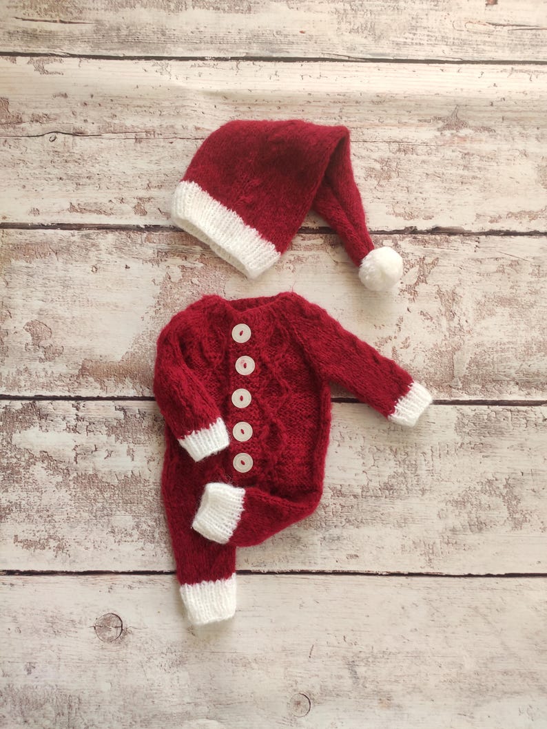 May include: Red knitted baby outfit with white trim and buttons, and a matching red knitted Santa hat with a white pom-pom.