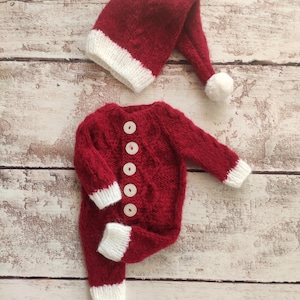 May include: Red knitted baby outfit with white trim and buttons. The outfit includes a matching hat with a white pom-pom.