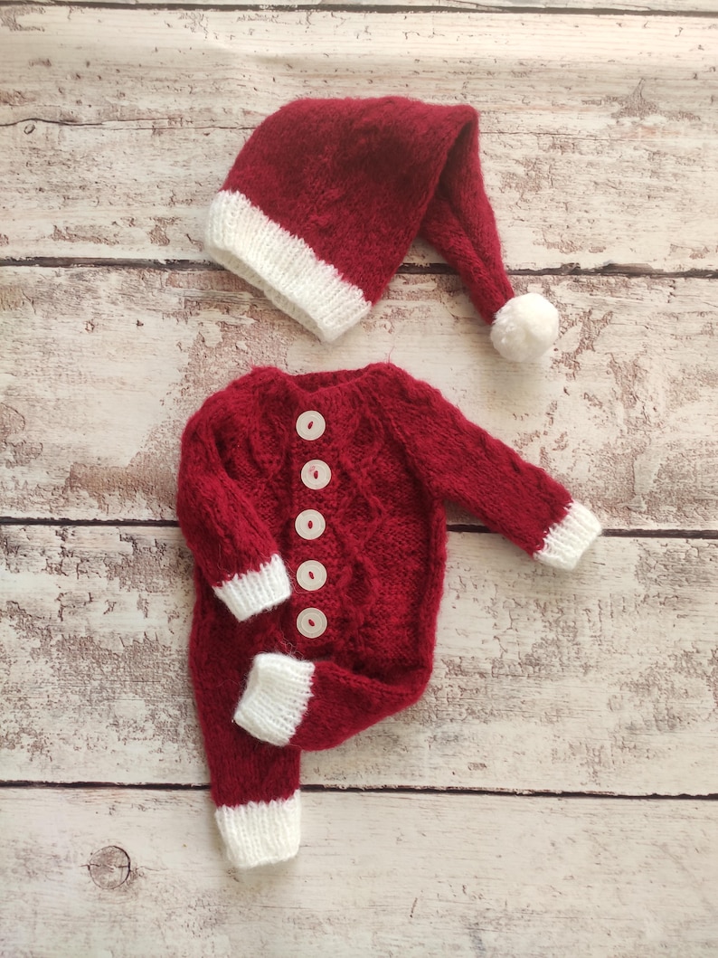 May include: Red knitted baby outfit with white trim and buttons. The outfit includes a matching hat with a white pom-pom.