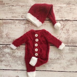 May include: Red knitted baby outfit with white trim and buttons. The outfit includes a matching Santa hat with a white pom-pom.