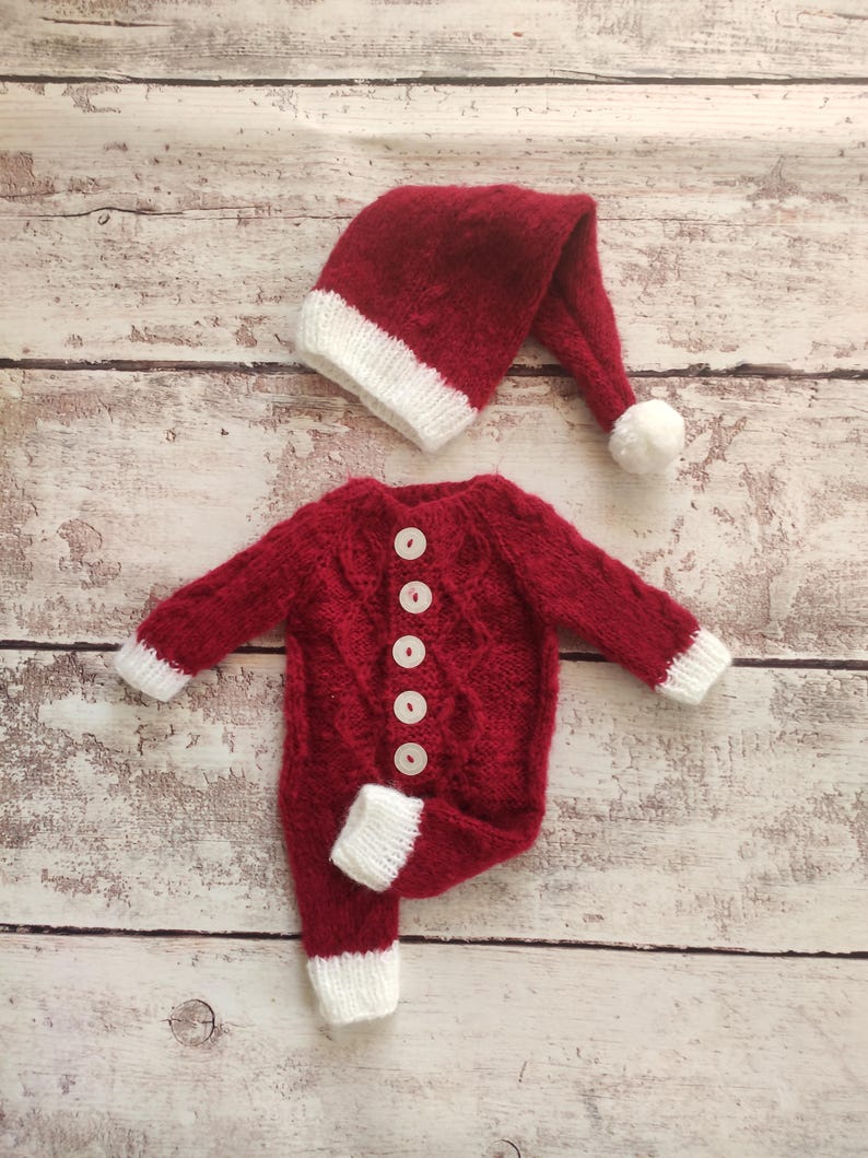 May include: Red knitted baby outfit with white trim and buttons. The outfit includes a matching Santa hat with a white pom-pom.