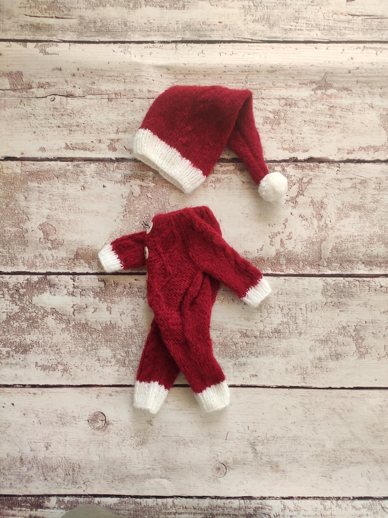 May include: Red knitted Santa suit with white trim for a newborn baby. The suit includes a matching hat with a white pom-pom.