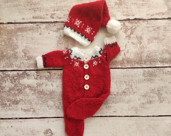 May include: Red knitted baby outfit with a white trim and snowflake pattern. The outfit includes a matching hat with a white pom-pom.