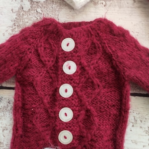 May include: A red knitted baby sweater with white trim and five white buttons down the front.
