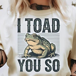 May include: A cream-coloured t-shirt with a detailed illustration of a green and tan toad. The text "I TOAD YOU SO" is printed above and below the toad graphic in a bold, dark font. The shirt is a casual wear item.