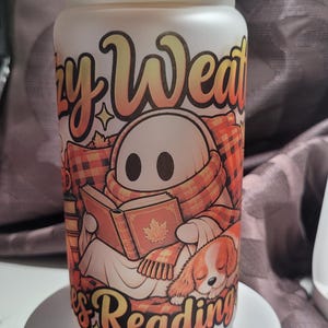 May include: A frosted glass tumbler with a bamboo lid and a clear straw. The exterior features a graphic of a ghost wearing a plaid scarf, reading a book with a maple leaf on the cover, next to a sleeping dog. The text reads Cozy Weather and Reading.