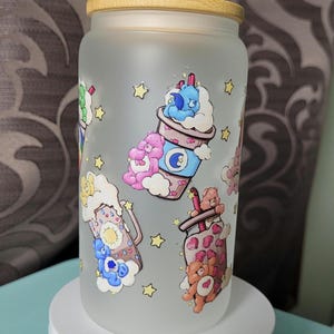 May include: A frosted glass tumbler with a bamboo lid featuring a repeating pattern of colourful cartoon bears, clouds, and stars. The design includes illustrations of bears interacting with beverage cups, decorated with symbols like a moon and sun.