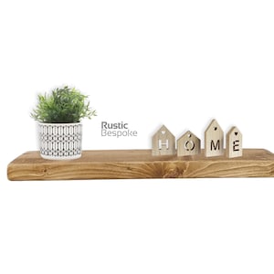 May include: A rectangular wooden shelf with a natural grain finish. On the left sits a white pot with a black geometric pattern containing a green plant. To the right are four small wooden house-shaped blocks spelling the word HOME.