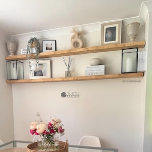 May include: Two medium brown floating shelves displaying decorative items. The shelves feature framed photos, vases, candles, books, and a plant. The wall is painted a neutral colour, creating a modern farmhouse style.