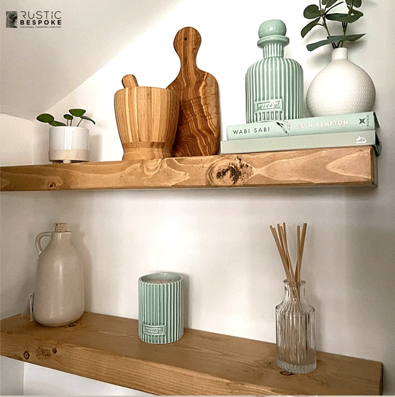 May include: Two wooden shelves display home decor items. The top shelf holds a mortar and pestle, a cutting board, a green bottle, a white vase, and books. The bottom shelf features a jug, a candle, and a glass diffuser with reeds.