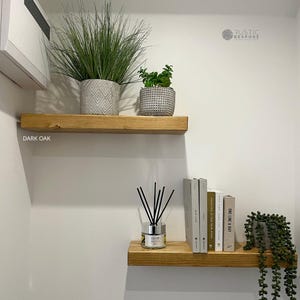 May include: Two dark oak floating shelves on a white wall. The top shelf displays a tall green plant in a grey patterned pot and a smaller green plant in a grey pot. The bottom shelf holds books, a reed diffuser, and a trailing plant.