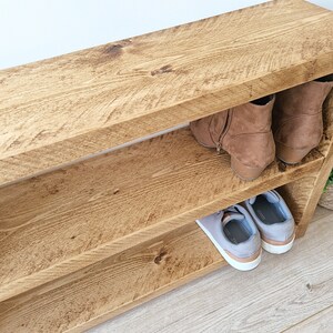 May include: A wooden shoe rack with two shelves. The top shelf has a pair of brown boots on it, and the bottom shelf has a pair of grey trainers.