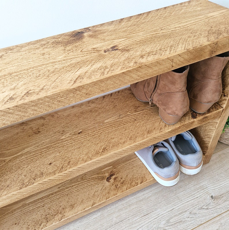 May include: A wooden shoe rack with two shelves. The top shelf has a pair of brown boots on it, and the bottom shelf has a pair of grey trainers.