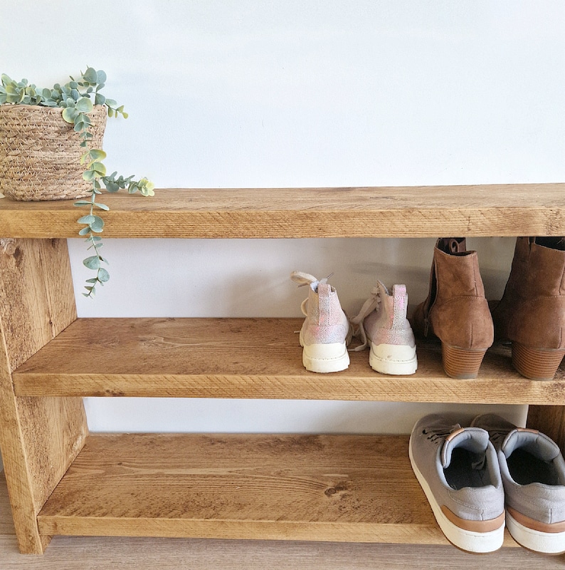 May include: A wooden shoe rack with two shelves. The top shelf has two pairs of pink and white trainers and two pairs of brown leather boots. The bottom shelf has two pairs of grey trainers.