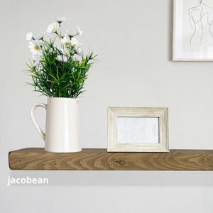 May include: A rustic wooden shelf displays a white pitcher with green and white flowers, a framed photo, and a minimalist artwork. The shelf is stained a warm brown color and is mounted on a white wall. The word "jacobean" is printed on the shelf.
