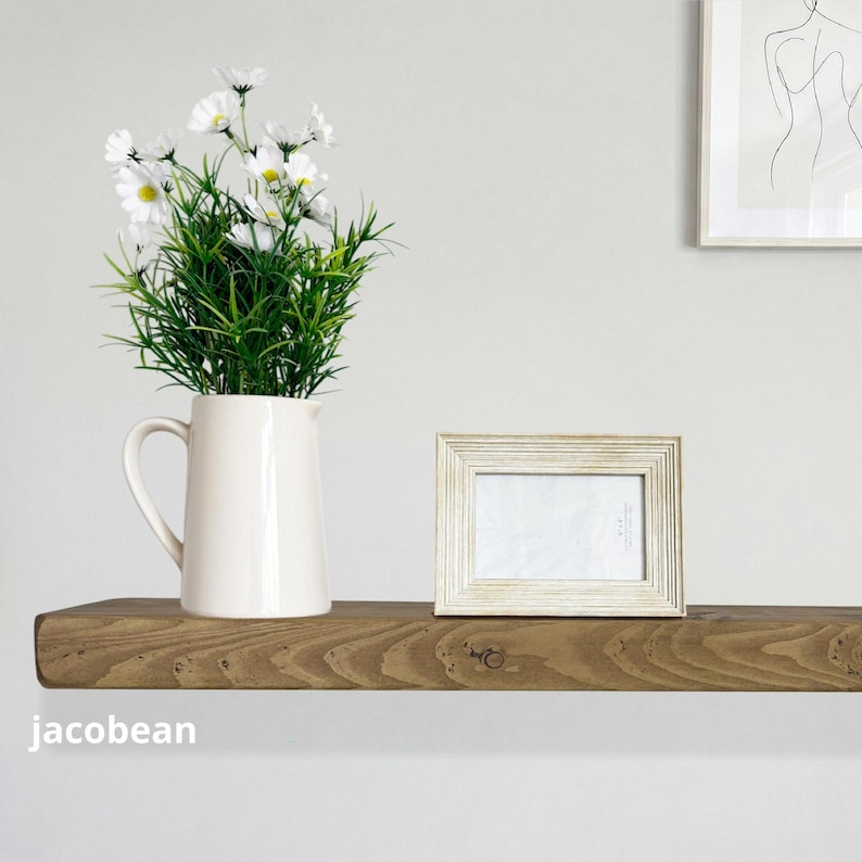 May include: A rustic wooden shelf displays a white pitcher with green and white flowers, a framed photo, and a minimalist artwork. The shelf is stained a warm brown color and is mounted on a white wall. The word "jacobean" is printed on the shelf.