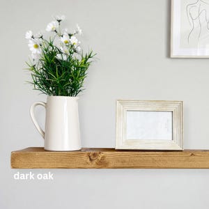 May include: A dark oak floating shelf displays a white pitcher with white flowers and a framed photo. The shelf is made of wood and has a knot in the wood. The text "dark oak" is visible on the shelf.