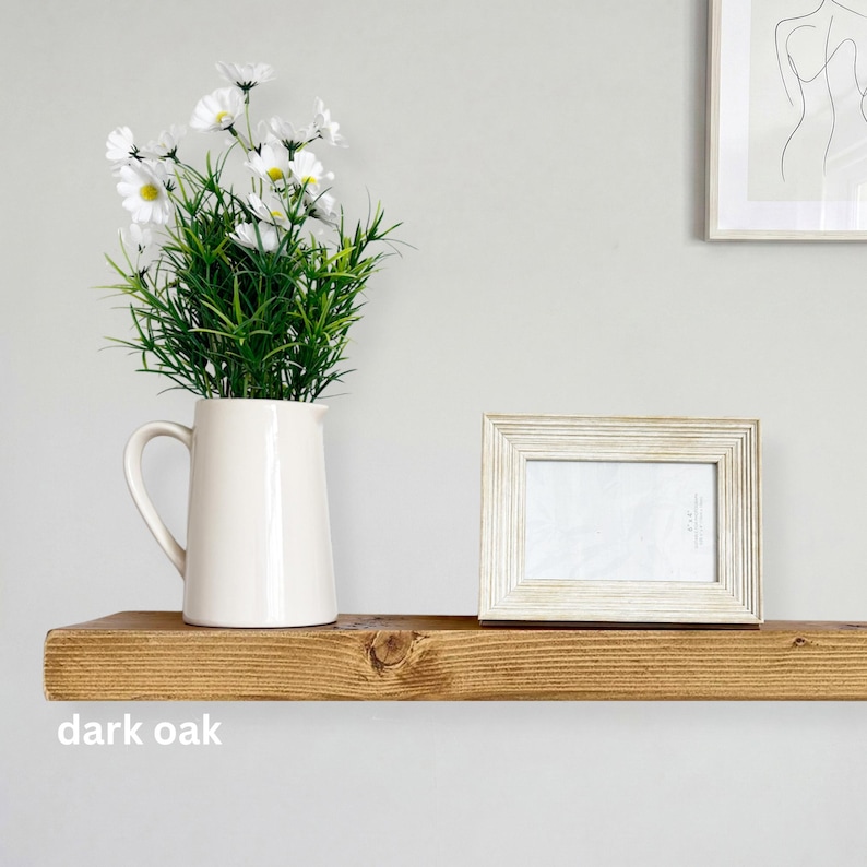 May include: A dark oak floating shelf displays a white pitcher with white flowers and a framed photo. The shelf is made of wood and has a knot in the wood. The text "dark oak" is visible on the shelf.