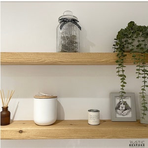 May include: A home decor scene featuring two wooden shelves against a white wall. The top shelf holds a glass jar with a black polka dot ribbon. The bottom shelf displays a white candle, a small tin, a framed dog portrait, and a brown diffuser bottle with reeds.