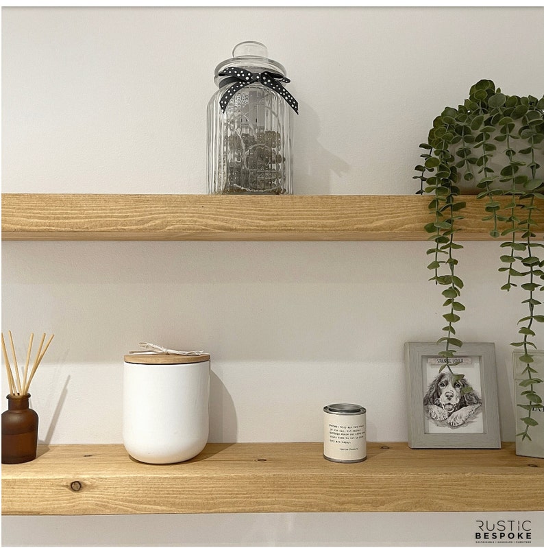 May include: A home decor scene featuring two wooden shelves against a white wall. The top shelf holds a glass jar with a black polka dot ribbon. The bottom shelf displays a white candle, a small tin, a framed dog portrait, and a brown diffuser bottle with reeds.
