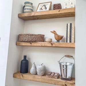 May include: Wooden shelves displaying various decorative items. A blue and white striped pitcher, framed artwork, a wooden die, and a small white house are on the top shelf. The middle shelf holds a woven basket, a small bowl, and a wooden duck. The bottom shelf has a black bottle, pear and apple sculptures, and a metal bucket.