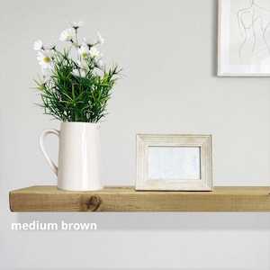 May include: A cream-colored pitcher with a handle holds a bouquet of white daisies and green foliage. A small, rectangular, light-colored picture frame sits next to the pitcher on a medium brown wooden shelf. The text "medium brown" is below the shelf.
