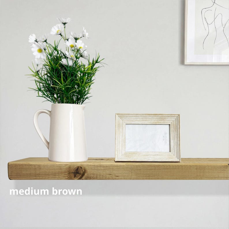 May include: A cream-colored pitcher with a handle holds a bouquet of white daisies and green foliage. A small, rectangular, light-colored picture frame sits next to the pitcher on a medium brown wooden shelf. The text "medium brown" is below the shelf.