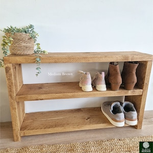 May include: A two-tiered wooden shoe rack in medium brown with two pairs of shoes on each shelf. The top shelf has a pair of brown ankle boots and a pair of pink trainers. The bottom shelf has a pair of grey trainers.