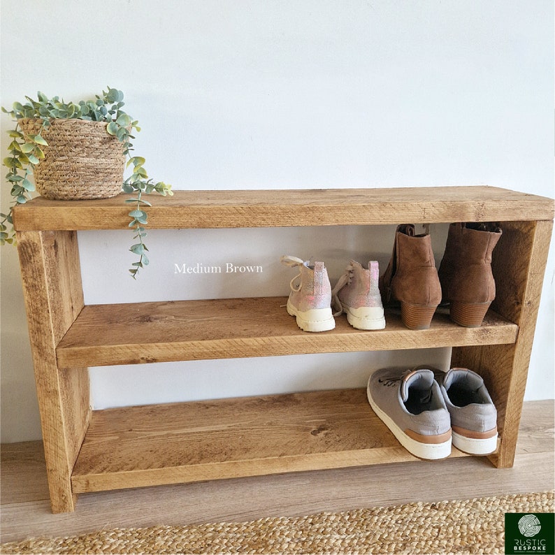 May include: A two-tiered wooden shoe rack in medium brown with two pairs of shoes on each shelf. The top shelf has a pair of brown ankle boots and a pair of pink trainers. The bottom shelf has a pair of grey trainers.