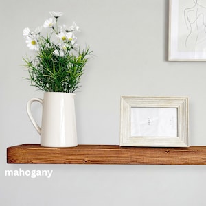 May include: A mahogany-colored wooden shelf with a white pitcher holding white daisies and green foliage. A white framed photo with the word "NEXT" is on the shelf. The word "mahogany" is at the bottom.