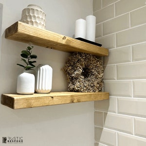 May include: Two rustic wooden floating shelves with decorative items. The top shelf holds white candles and a textured vase. The bottom shelf displays white vases, greenery, and a dried flower wreath. The shelves are mounted on a neutral-colored wall.