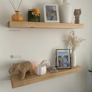 May include: Two medium brown wooden floating shelves on a white wall, each displaying decorative items. The top shelf features a pumpkin diffuser, a jar with a flower, a framed photo, a vase, and a figurine. The bottom shelf has an elephant figurine, a candle, a shell, a framed photo, and a vase.