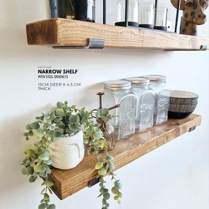 May include: Rustic wood shelf with steel brackets. The shelf is 15 inches deep and 4.5 inches thick. It is decorated with glass jars, a potted plant, and a small watering can.