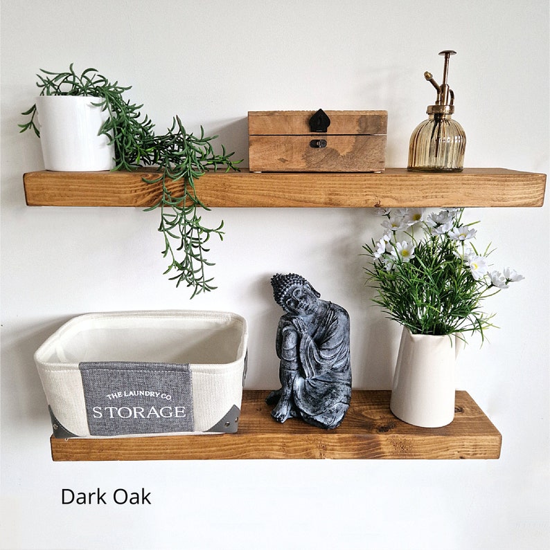 May include: Two dark oak floating shelves with decorative items. The top shelf holds a white potted plant, trailing greenery, a wooden box, and a glass spray bottle. The bottom shelf displays a storage basket, a Buddha statue, and a vase of white flowers.
